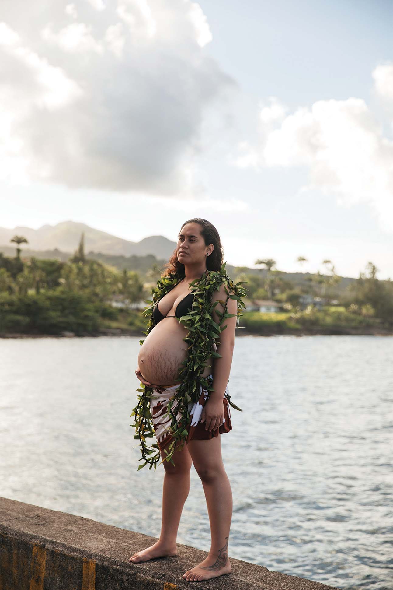 a pregnant person standing on a dock with a green garland around her belly, a photo by Nani Welch Keli‘iho‘omalu