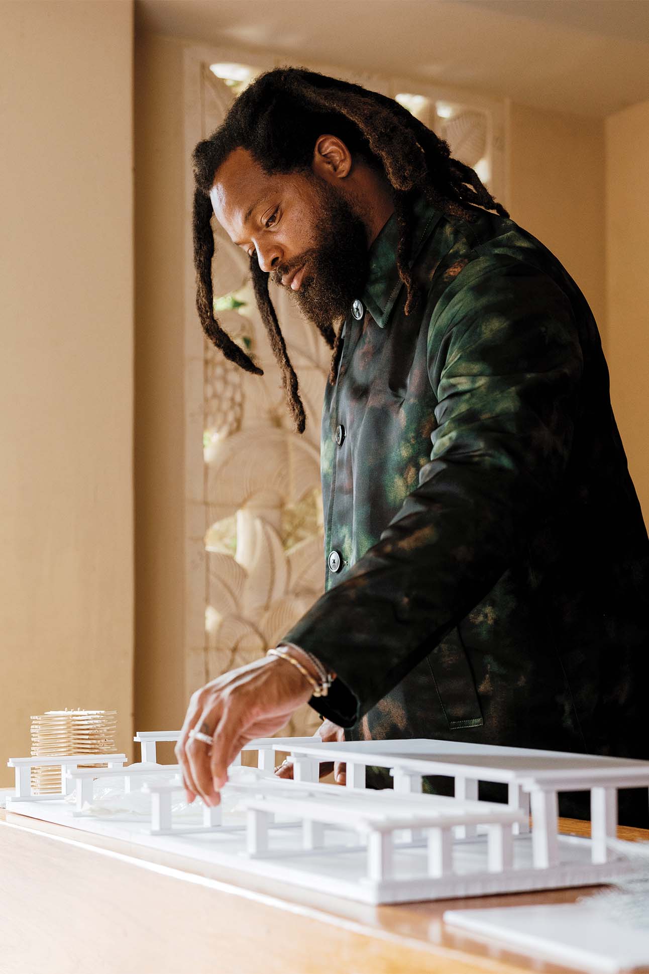 A man with dreadlocks examines and adjusts a white architectural model of a building placed on a table, wearing a dark green camo-style shirt.