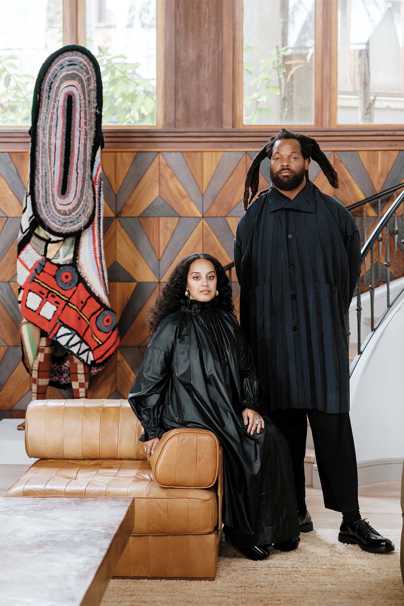 A woman seated on a tan leather bench and a man standing next to her in a room with wooden geometric wall panels and an abstract colorful textile sculpture in the background. Both are dressed in elegant black attire.