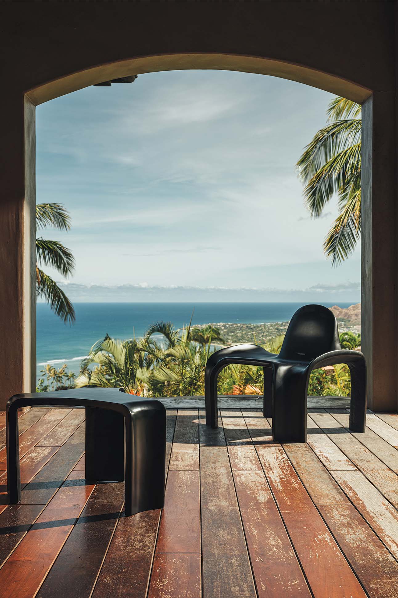 View from a large arched doorway showing a wooden deck with two modern black chairs overlooking a lush coastal landscape with palm trees, blue sea and distant mountains.