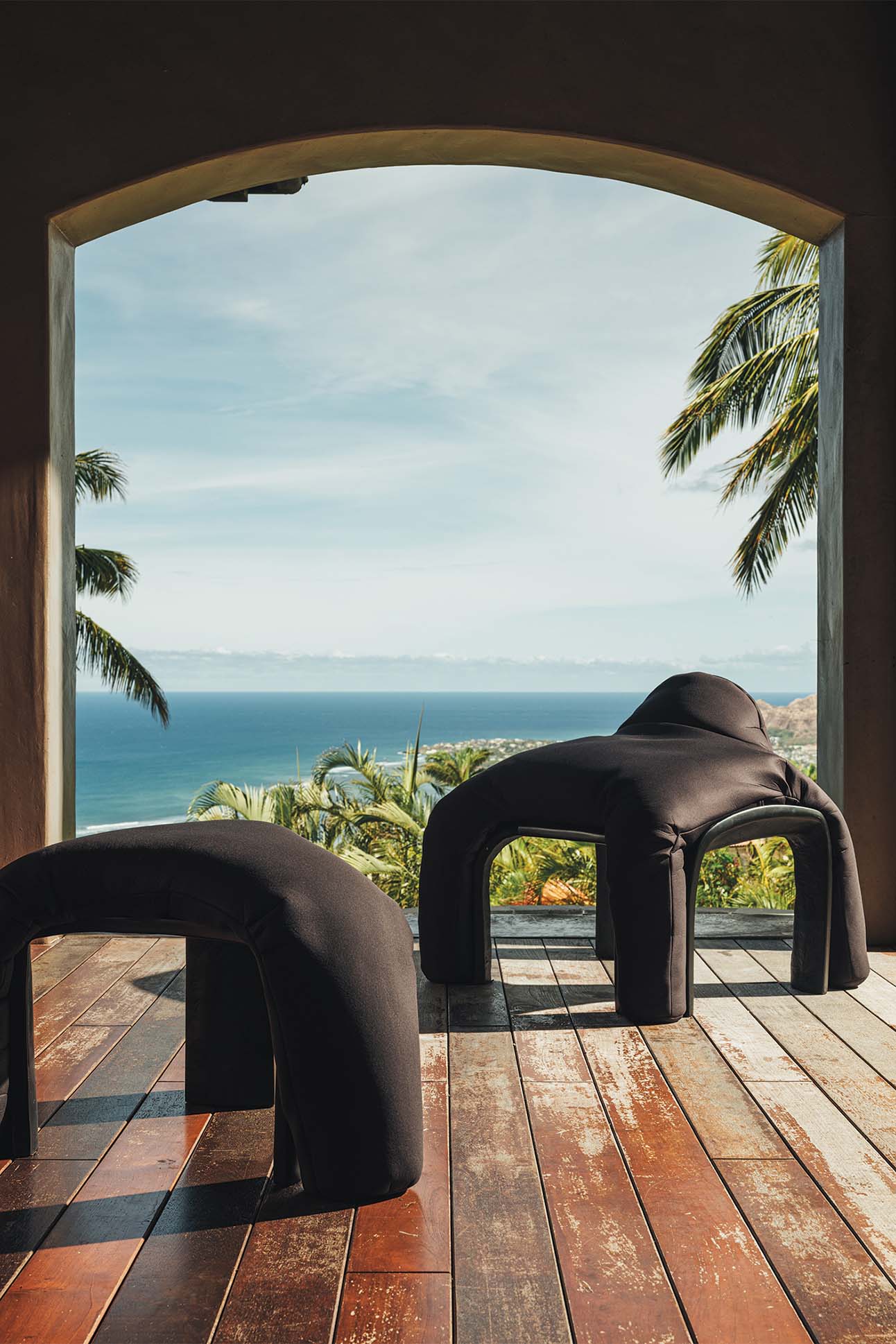 View through an archway featuring two modern black chairs on a wooden floor overlooking a tropical landscape with palm trees and a distant ocean under a clear blue sky.